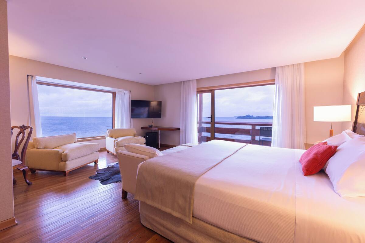 A hotel room with a large bed, two armchairs, a TV, and a view of the sea.