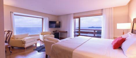 Habitación de hotel con una cama grande, dos sillones, un televisor y vista al mar.