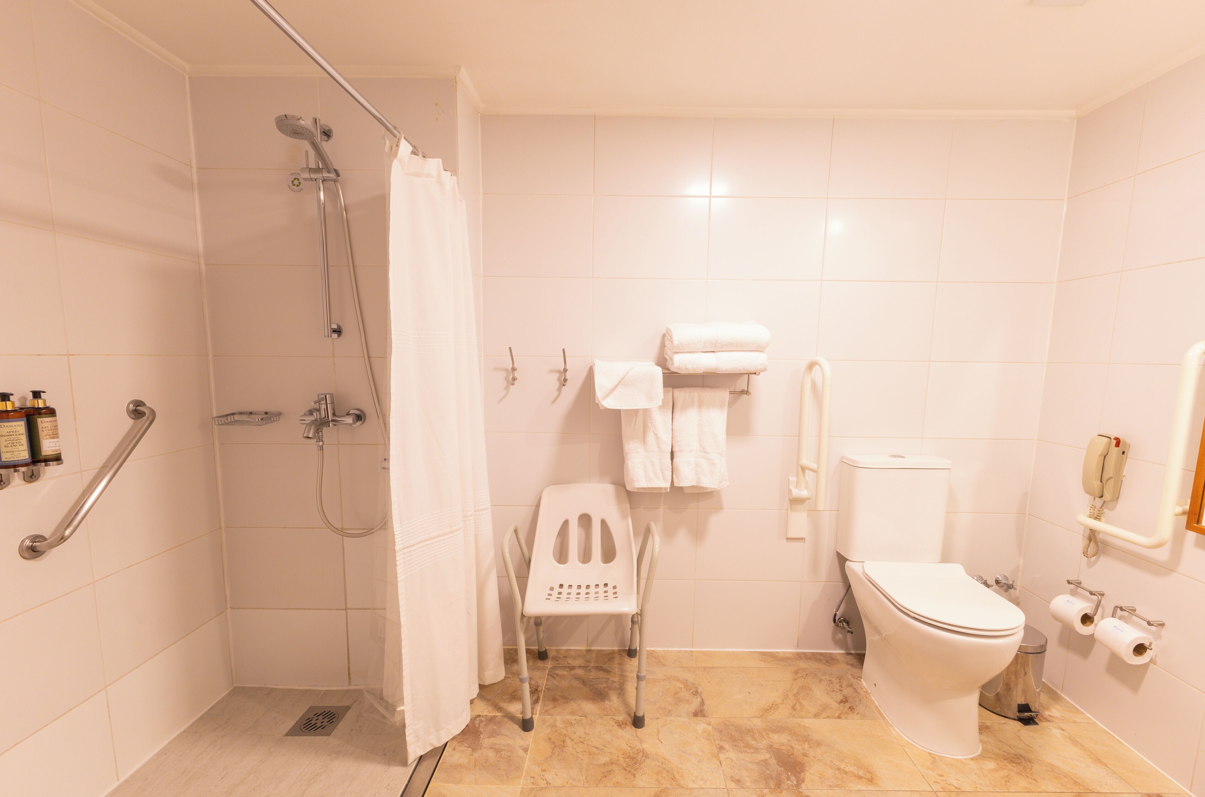 A bathroom with a shower, toilet, grab bars, and a wheelchair-accessible chair.