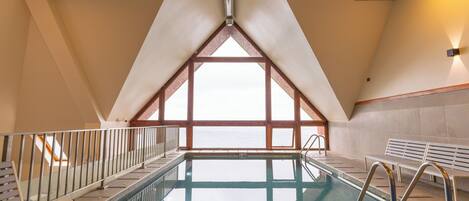 An indoor swimming pool with a view of the sea, a wooden deck, and a high ceiling.
