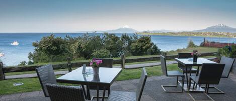 An outdoor patio with tables and chairs, overlooking a body of water and a mountain.