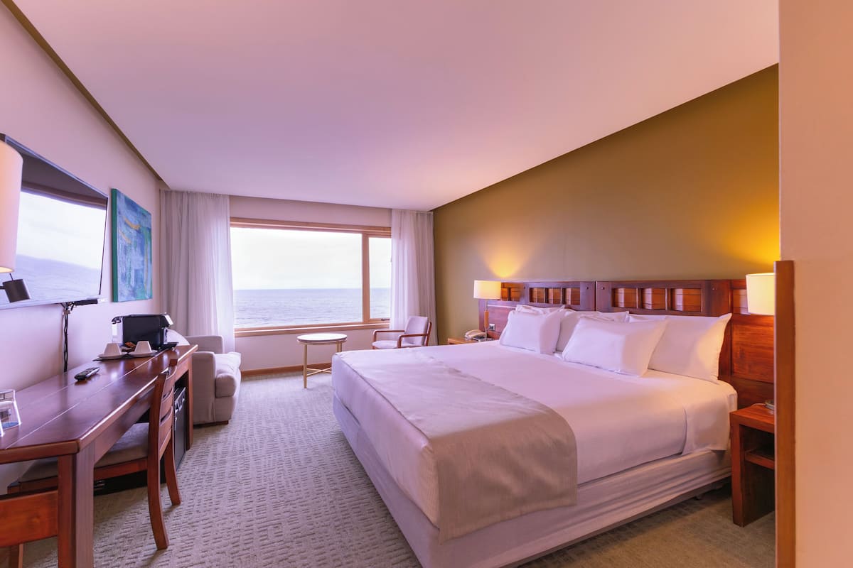 A hotel room with a bed, desk, chair, and a view of the ocean.