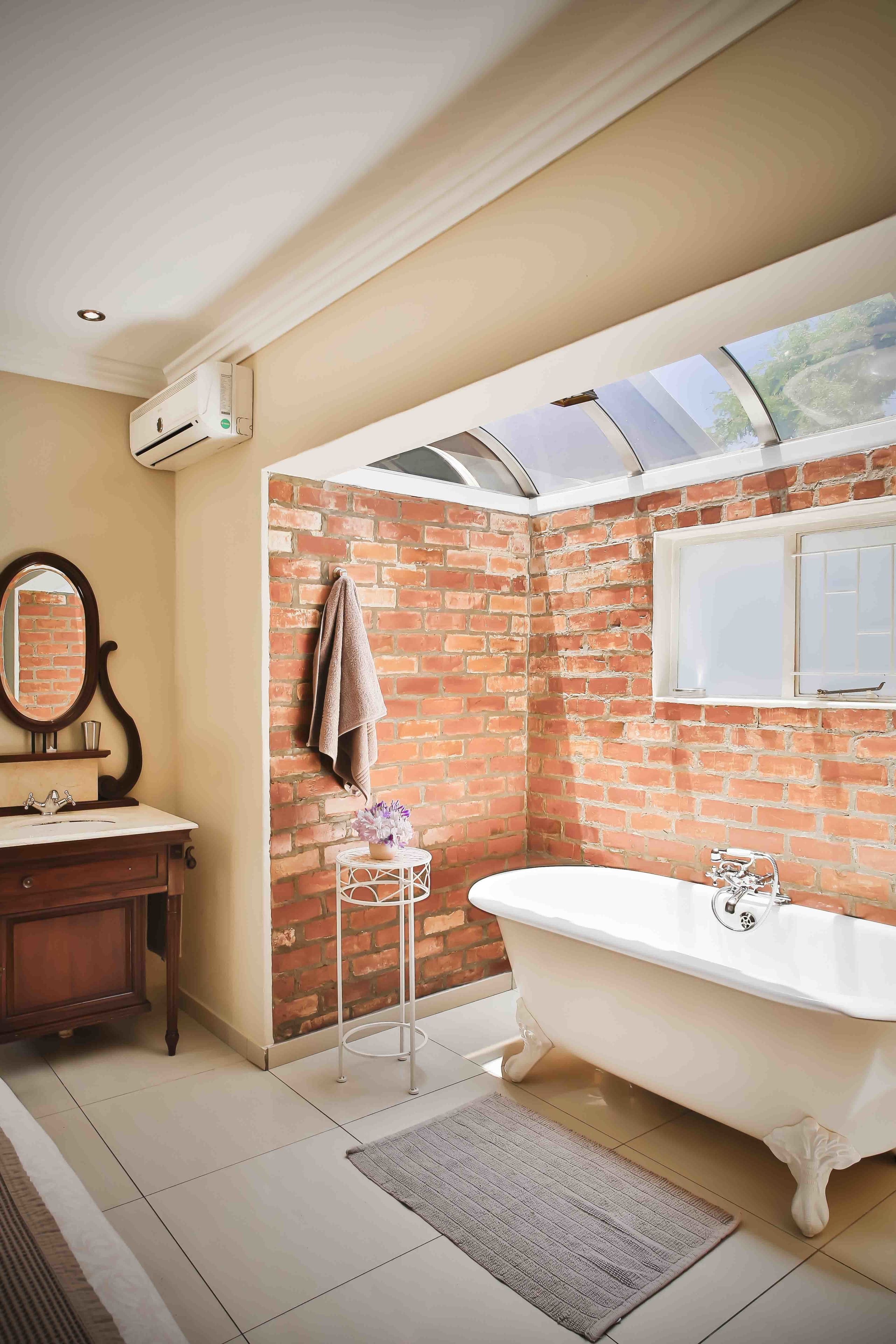 Un baño con pared de ladrillo, una bañera blanca, tocador de madera y un espejo grande.