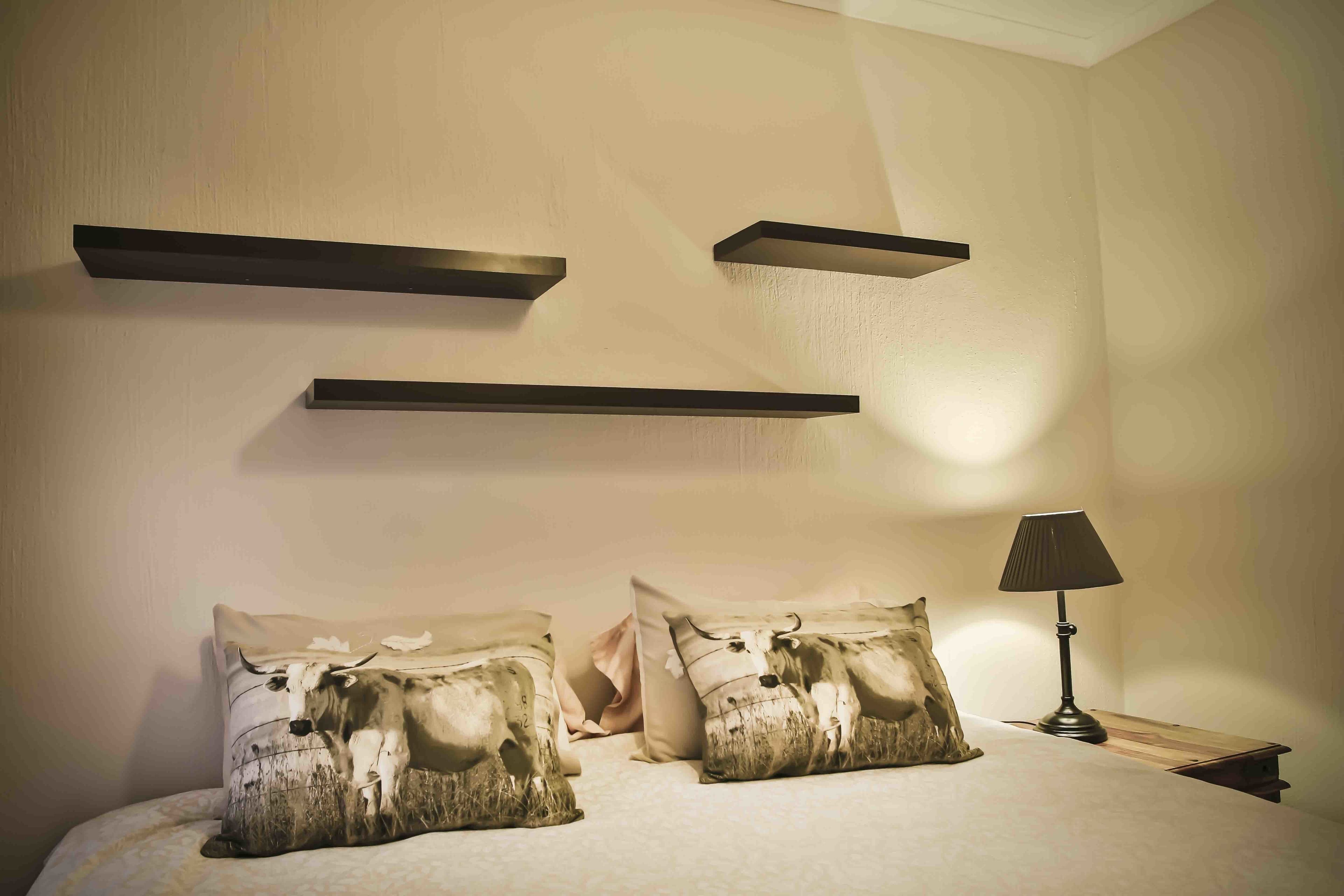A bedroom with a bed, two pillows with a cow print design, a bedside table with a lamp, and three wall-mounted shelves.