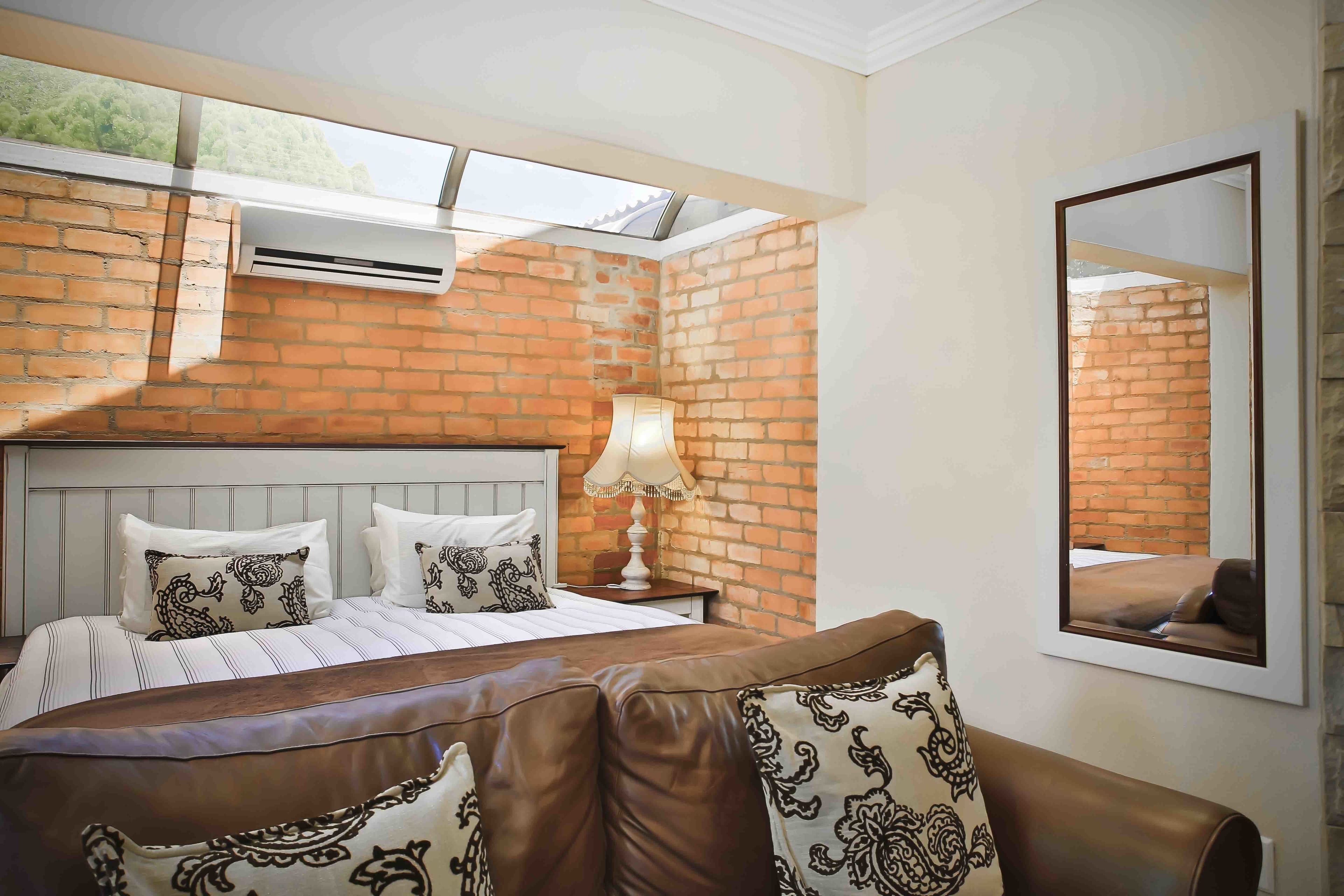 A bedroom with a brick wall, a bed with striped bedding, a brown leather sofa, a nightstand with a lamp, and a mirror on the wall.