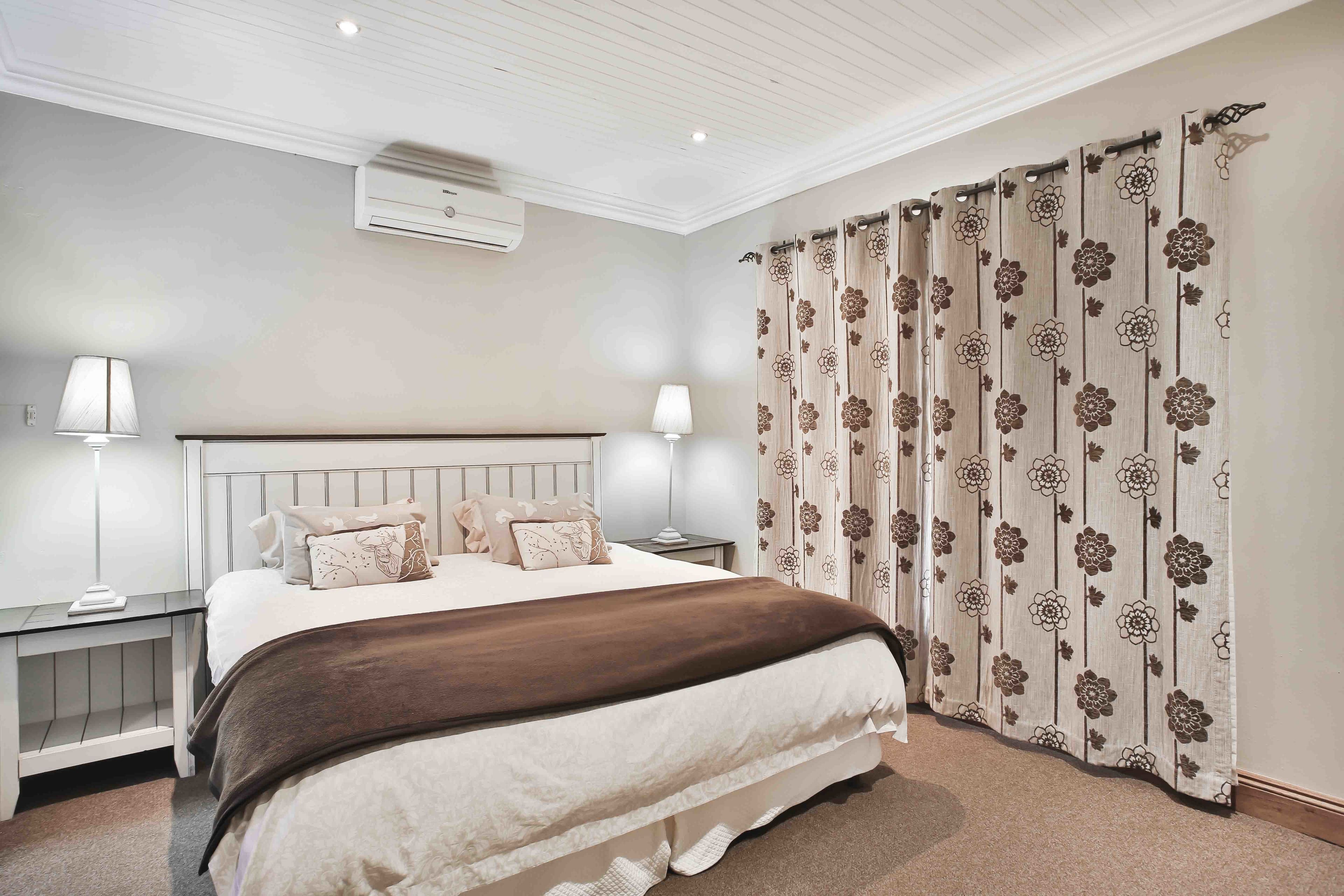 Un dormitorio con una cama grande, dos mesitas de noche, un buró, aire acondicionado instalado en la pared y una cortina con estampado floral.