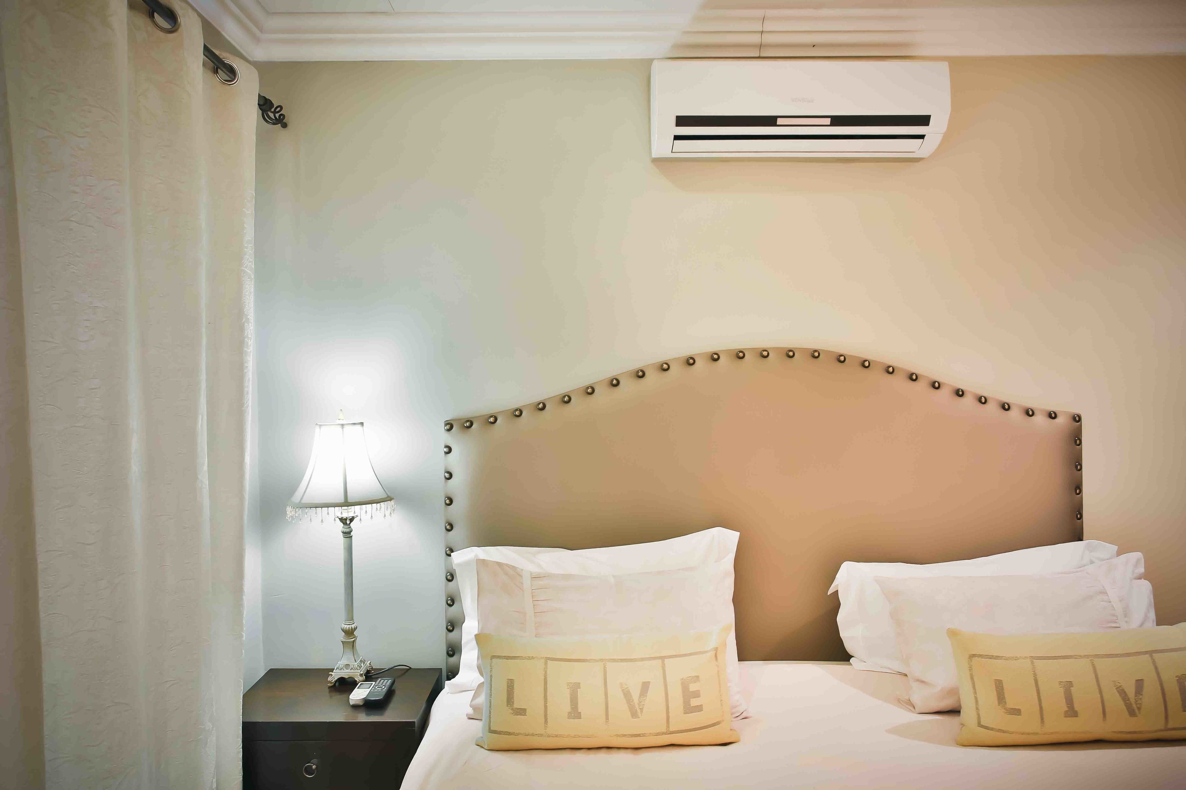Una cama bien hecha con dos almohadas, una lámpara de noche y un equipo de aire acondicionado en la pared.