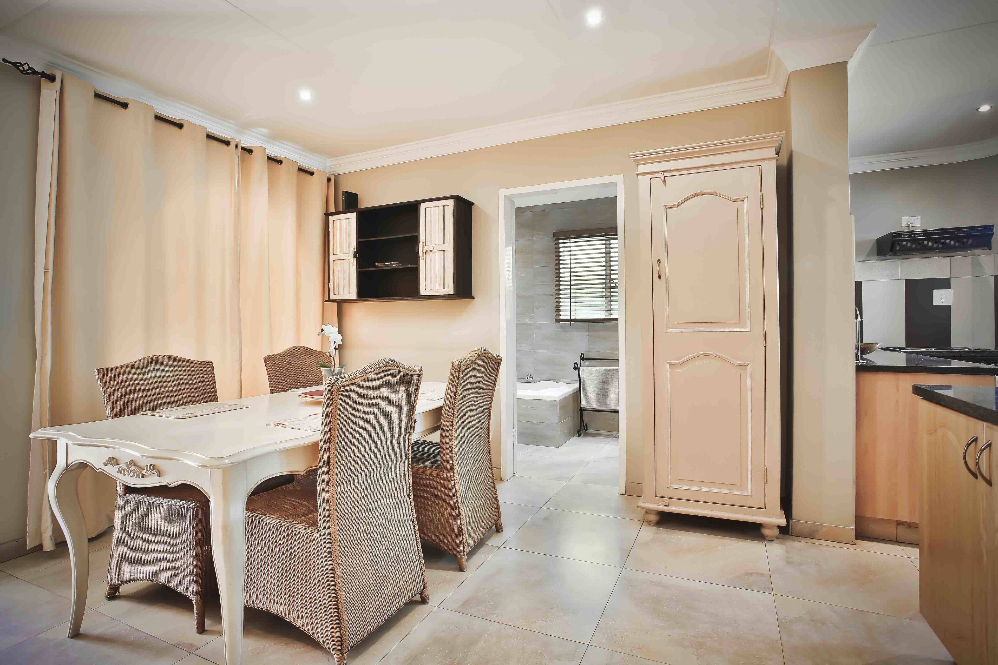 Un comedor con una mesa blanca y sillas de mimbre, una cocina con armarios de madera y un baño visible a través de un pasillo abierto.