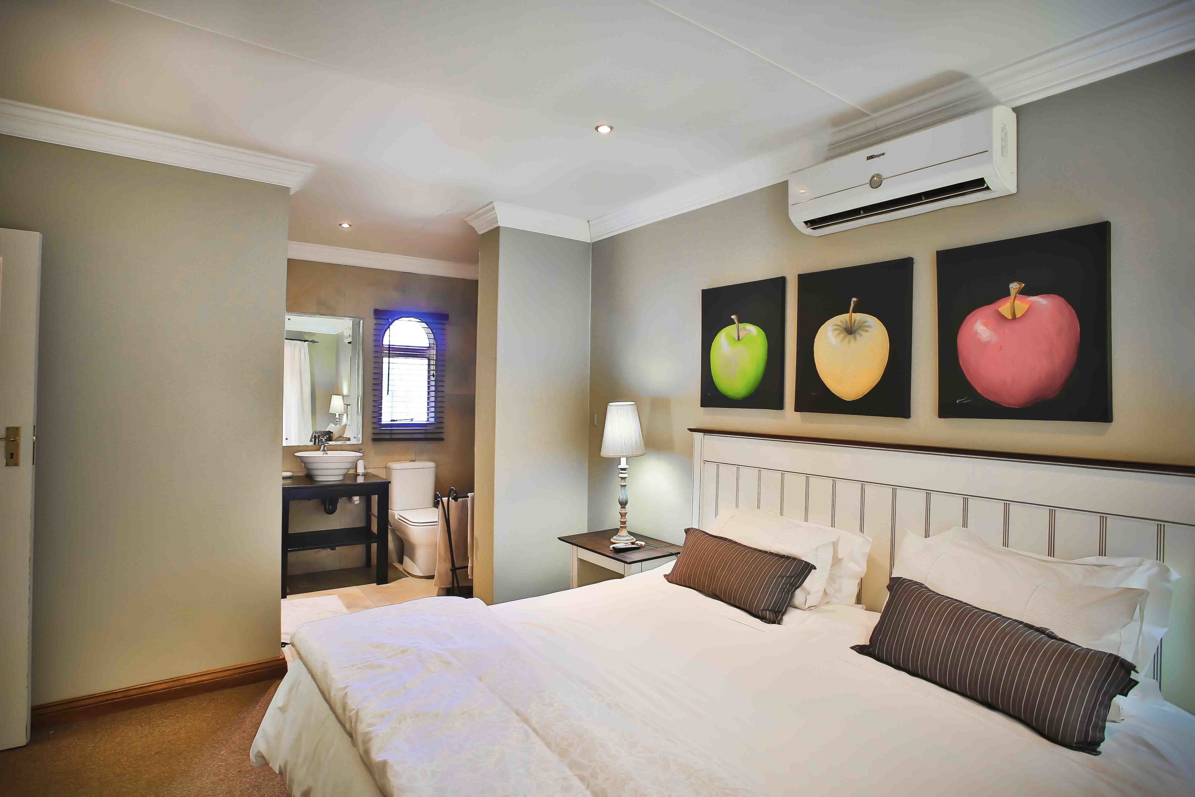 A hotel room with a large bed, a nightstand with a lamp, a wall-mounted air conditioner, and three framed fruit paintings.