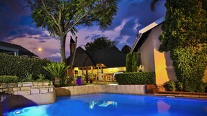 Una piscina con borde de piedra, rodeada de vegetación y un edificio con techo de paja.
