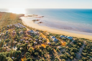 An aerial view of a coastal resort with multiple bungalow-style accommodations, a sandy beach, and a clear view of the ocean.