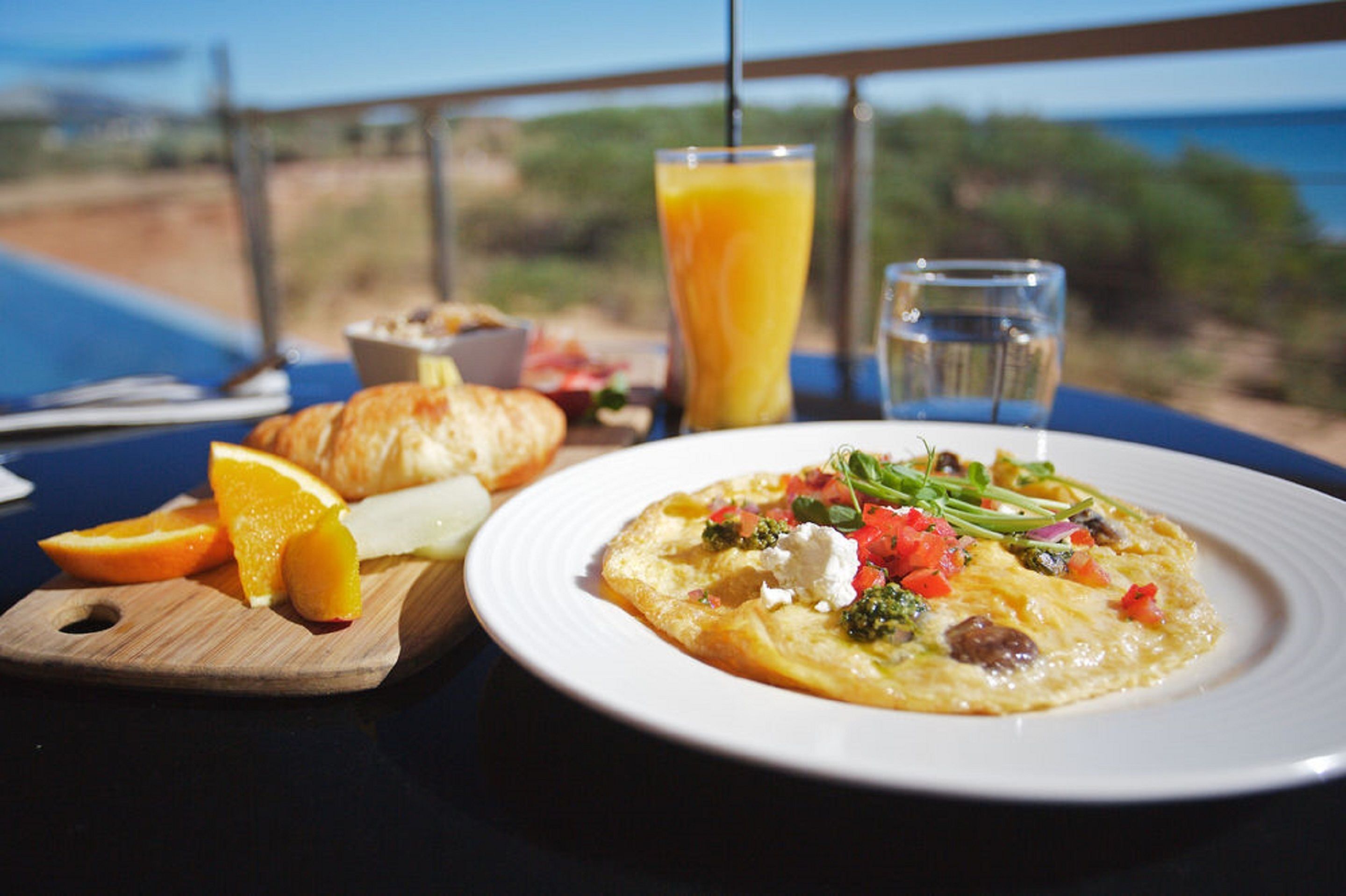 A breakfast setting with a plate of food, a glass of orange juice, and a water glass on a table with a view of the ocean.
