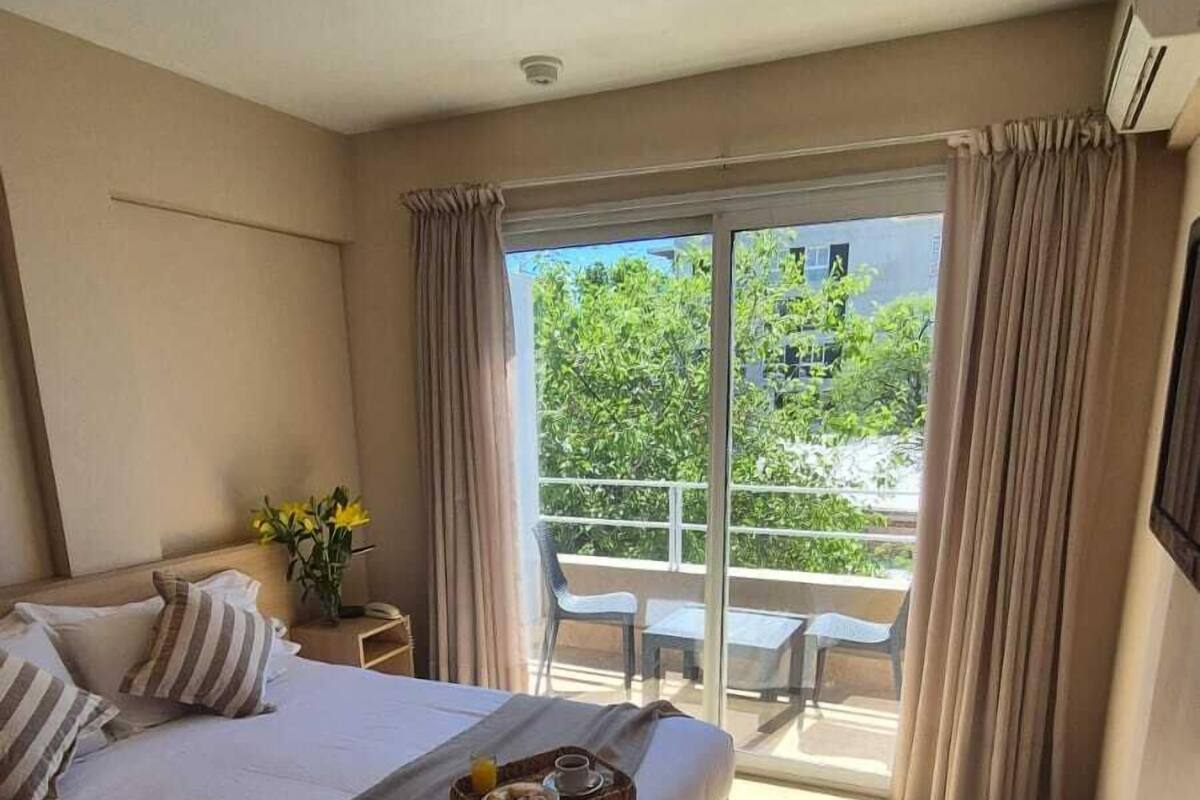 A hotel room with a bed, a small table with a vase of flowers, a balcony with a table and chairs, and a view of greenery outside.
