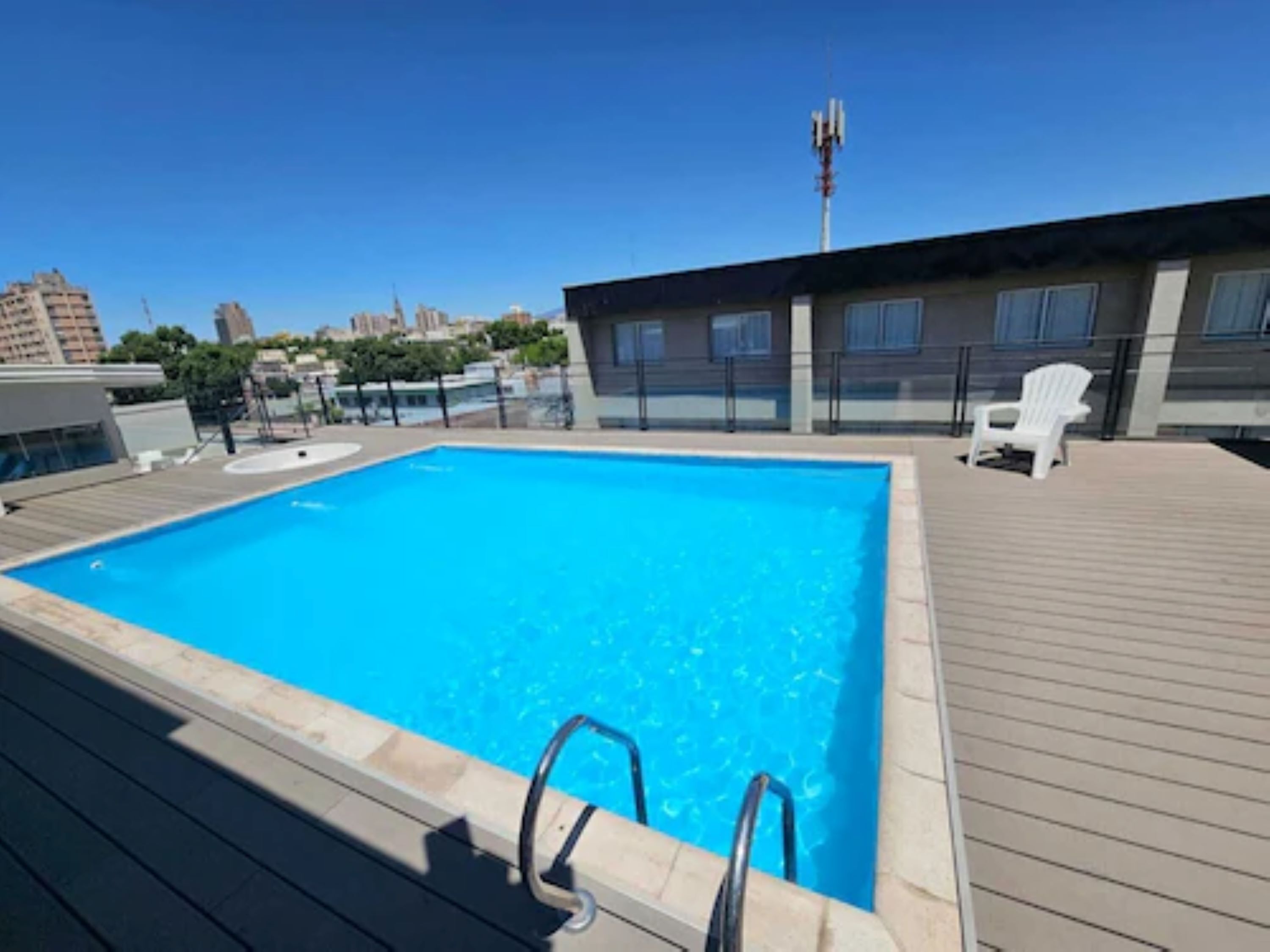 En pool på taket med ett trädäck, en vit stol och en stadsbild i bakgrunden.