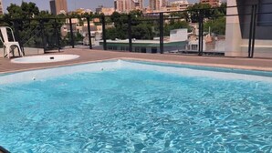 Una piscina en la azotea con agua azul cristalina, una escalera metálica y una vista panorámica de la ciudad al fondo.
