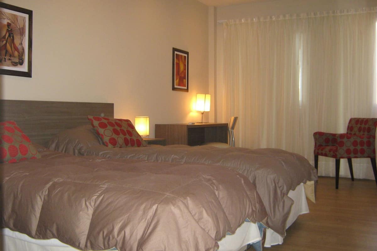 A double bed with a headboard, a bedside table with a lamp, a chair, and a painting on the wall.