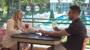 Two people seated at a table with drinks, overlooking a pool area with umbrellas and lounge chairs.
