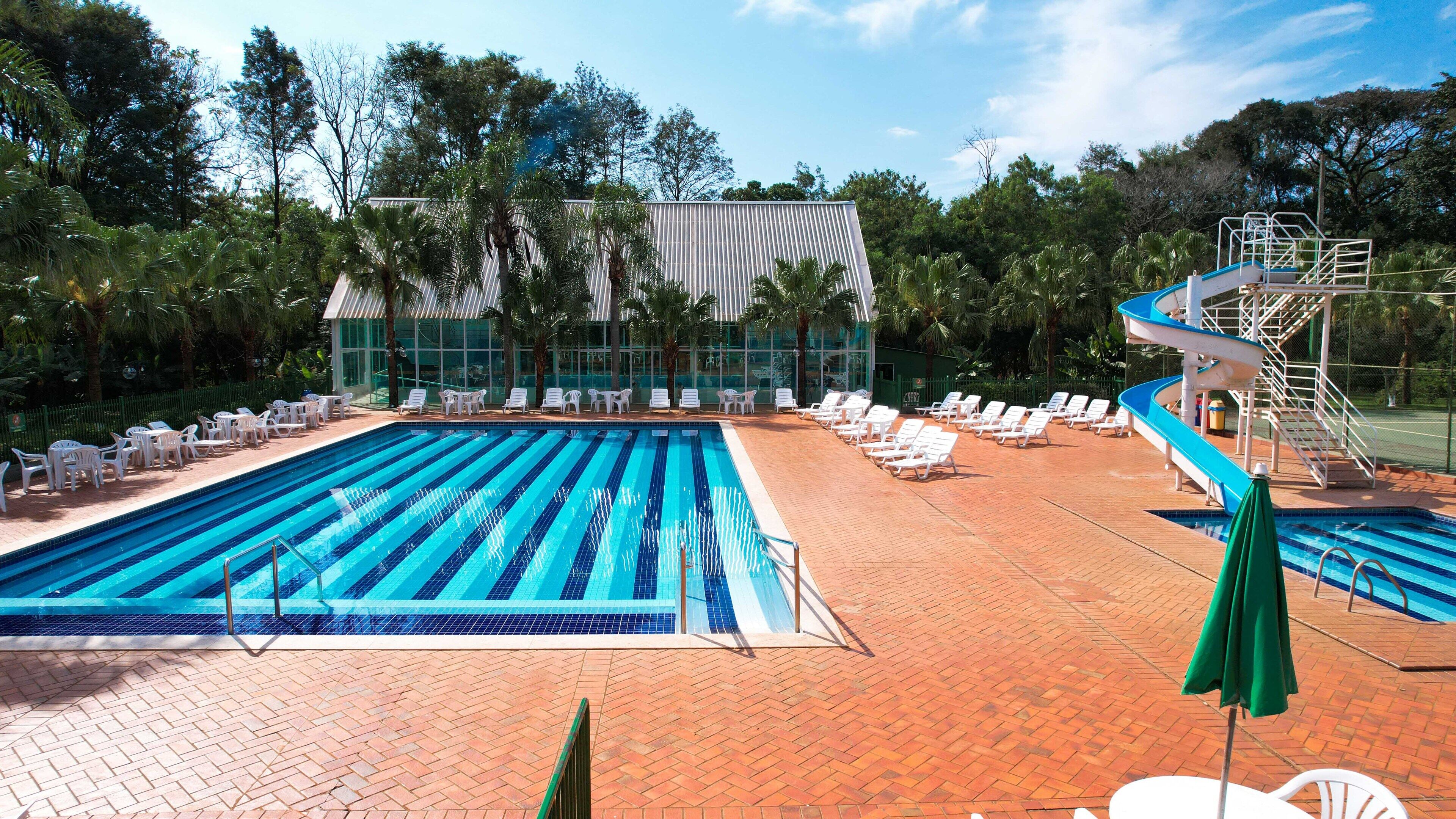 A swimming pool with lounge chairs and a slide, surrounded by trees and a building with a glass facade.