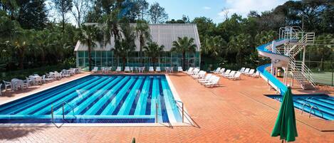 A swimming pool with lounge chairs and a slide, surrounded by trees and a building with a glass facade.