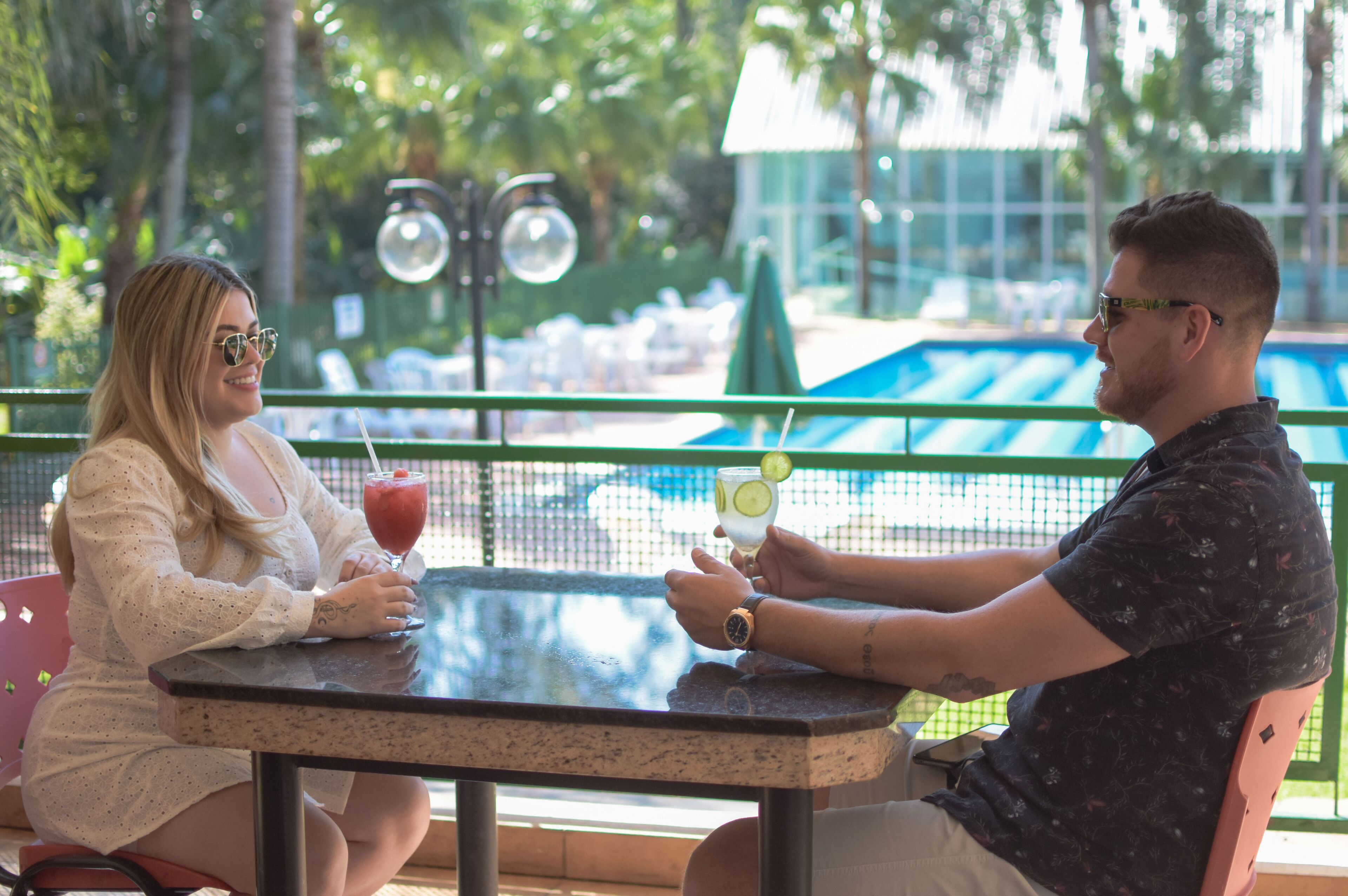 Two people seated at an outdoor table with drinks, overlooking a pool area.