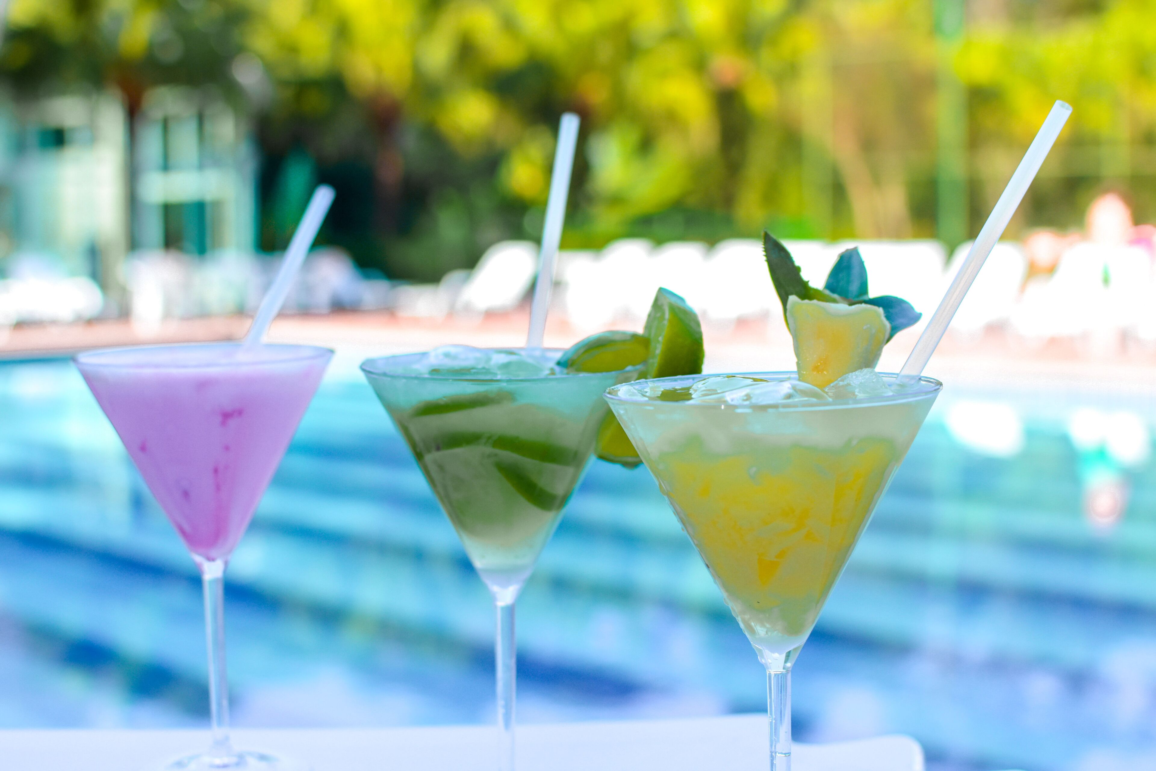 Drie cocktails met rietjes, versierd met limoen en ananas, op een tafel bij een zwembad.