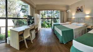 A hotel room with a desk, two beds, a TV, and large windows offering a view of greenery.