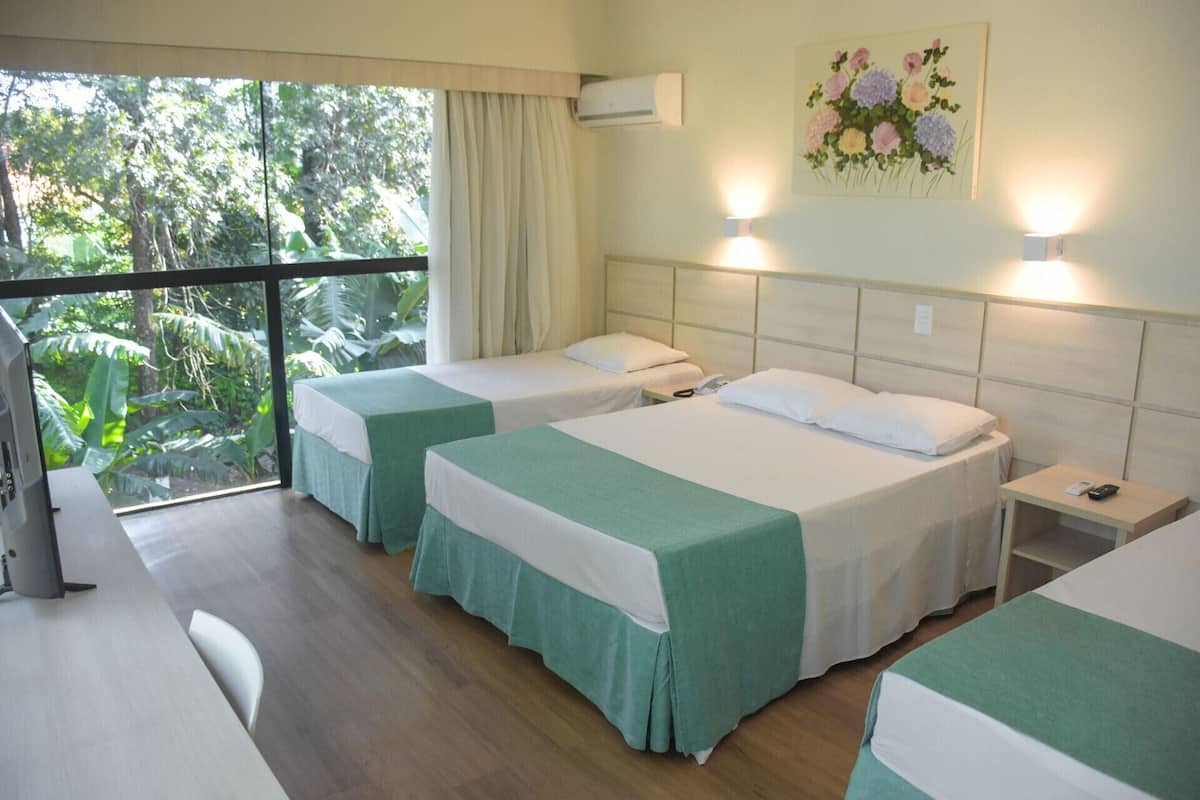 A hotel room with two beds, a desk, a chair, and a large window with a view of trees.