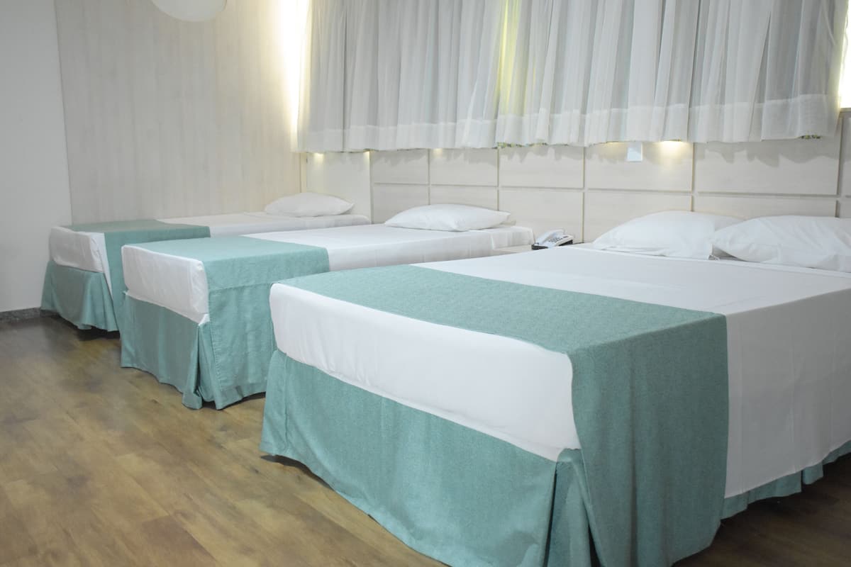 A hotel room with three single beds, each with a white and teal bedspread. The room has wooden flooring and white walls.
