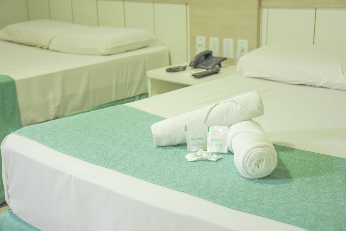 A hotel room with two beds, each with a green bedspread and white linens. There is a telephone on the nightstand and toiletries on the bed.