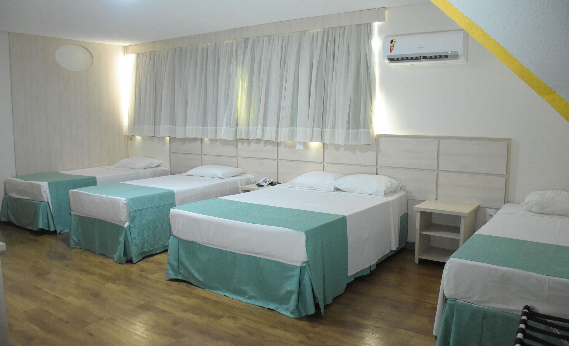 Une chambre d’hôtel avec quatre lits simples, une petite table de chevet avec un téléphone, un climatiseur fixé au mur et une fenêtre avec des rideaux légers.