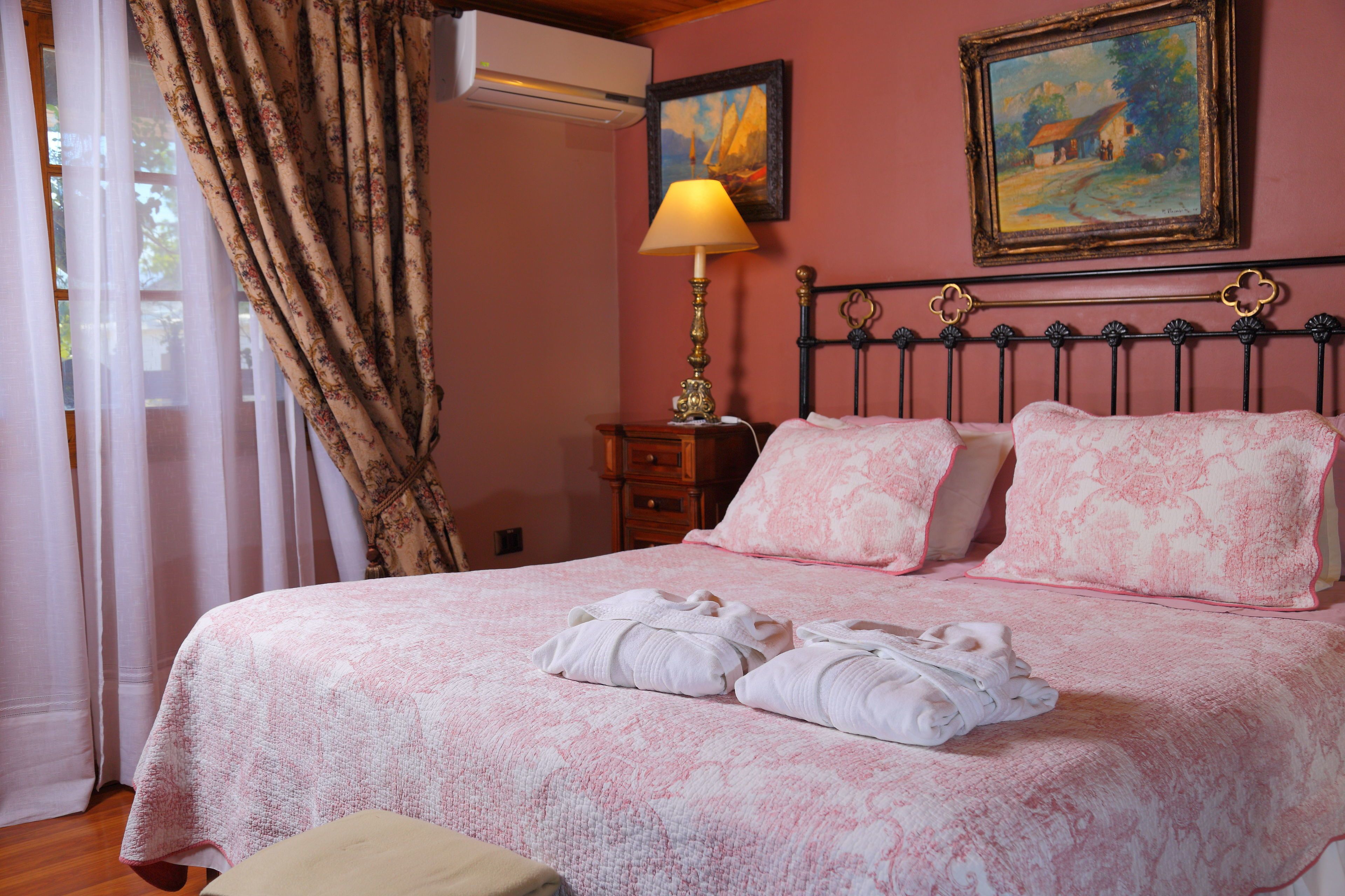 A neatly made bed with pink bedding, a lamp on a bedside table, and a painting on the wall.