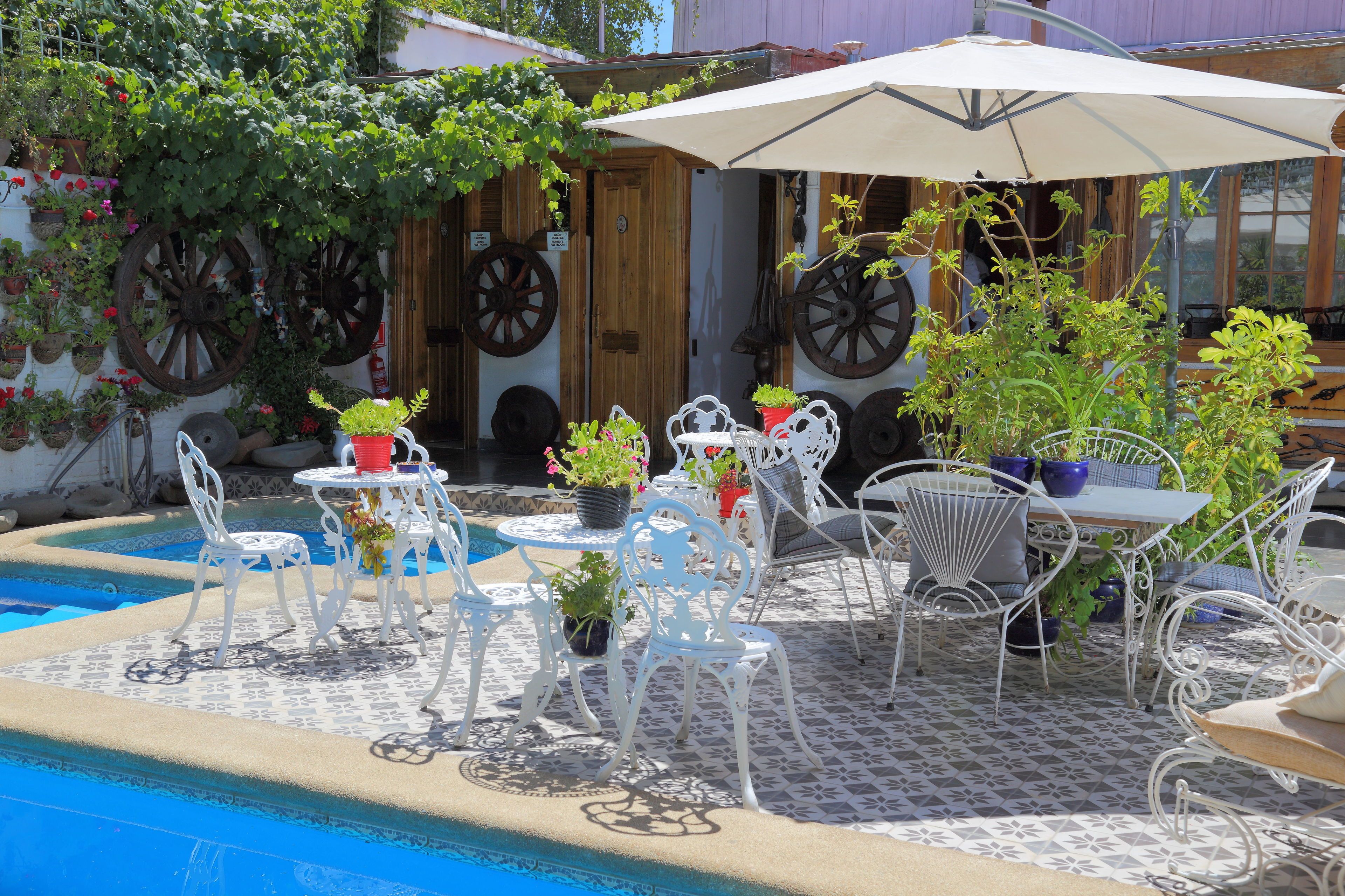 An outdoor patio with white metal chairs and tables, surrounded by potted plants and a pool.