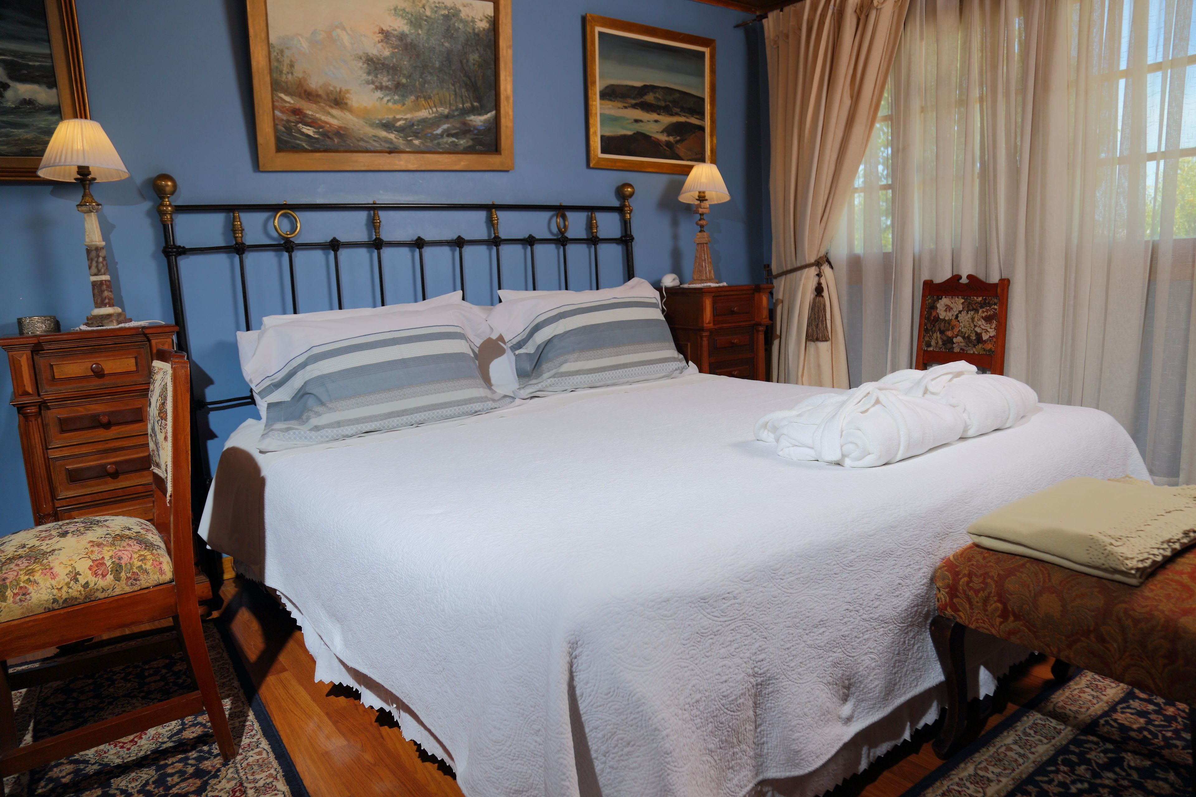 A neatly made bed with a white duvet and striped pillows, a wooden nightstand with a lamp, a chair with a floral cushion, and two framed paintings on the wall.