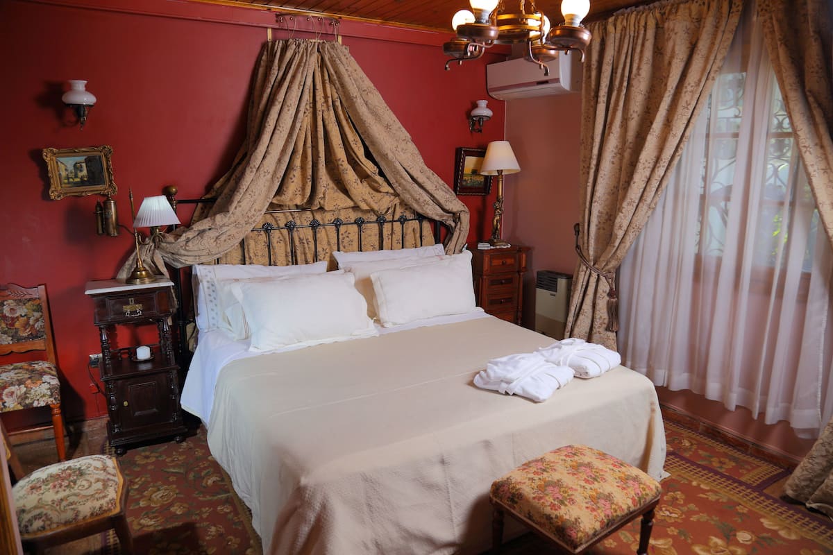 Una cama bien hecha con dosel, una mesita de noche de madera con lámpara, una silla con cojín floral y una pared roja con un marco de fotos.