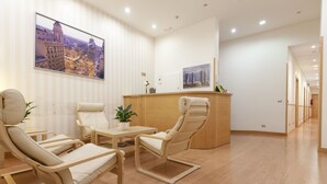 Un moderno vestíbulo de hotel con un mostrador de recepción, dos sillones, una pequeña mesa con una planta y un cuadro de gran formato con una vista urbana.