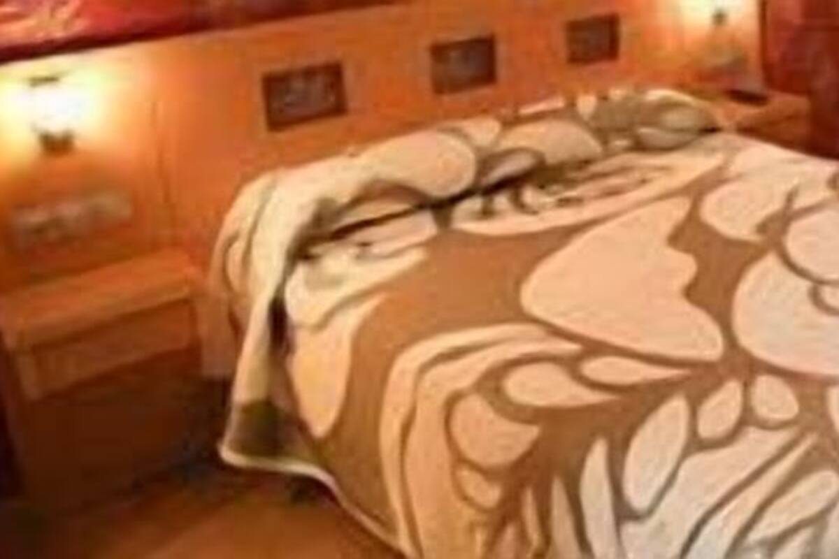 Quarto de hotel com uma cama coberta por um edredom estampado com motivos florais, uma mesa de cabeceira de madeira e uma luminária fixada na parede.