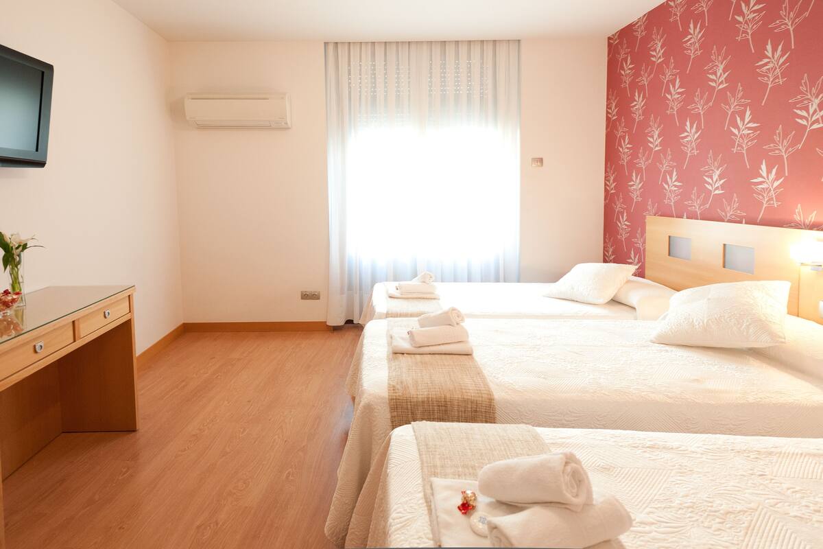 Quarto de hotel com duas camas, uma escrivaninha de madeira, televisão de tela plana e janela com cortinas translúcidas.