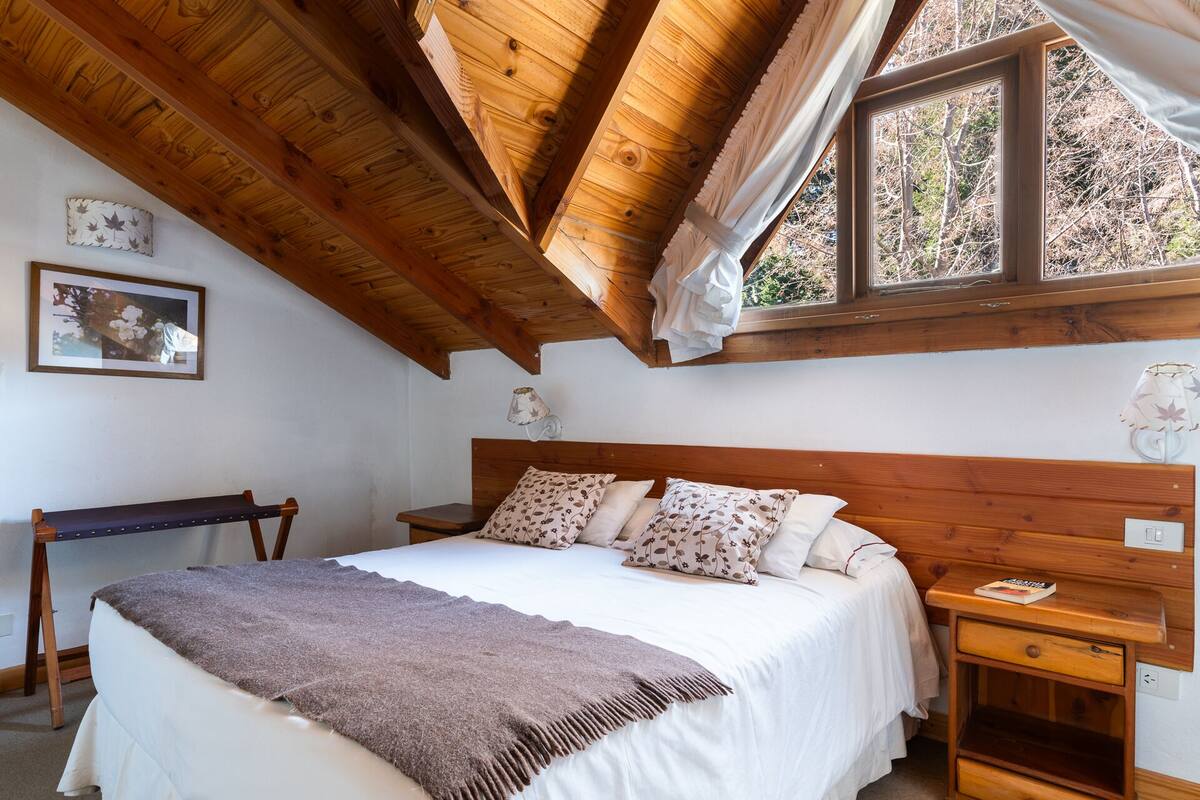 Ein Schlafzimmer mit Holzbalkendecke, einem Bett mit floralen Kissen, einer Bank und einem Fenster mit Blick auf Bäume.