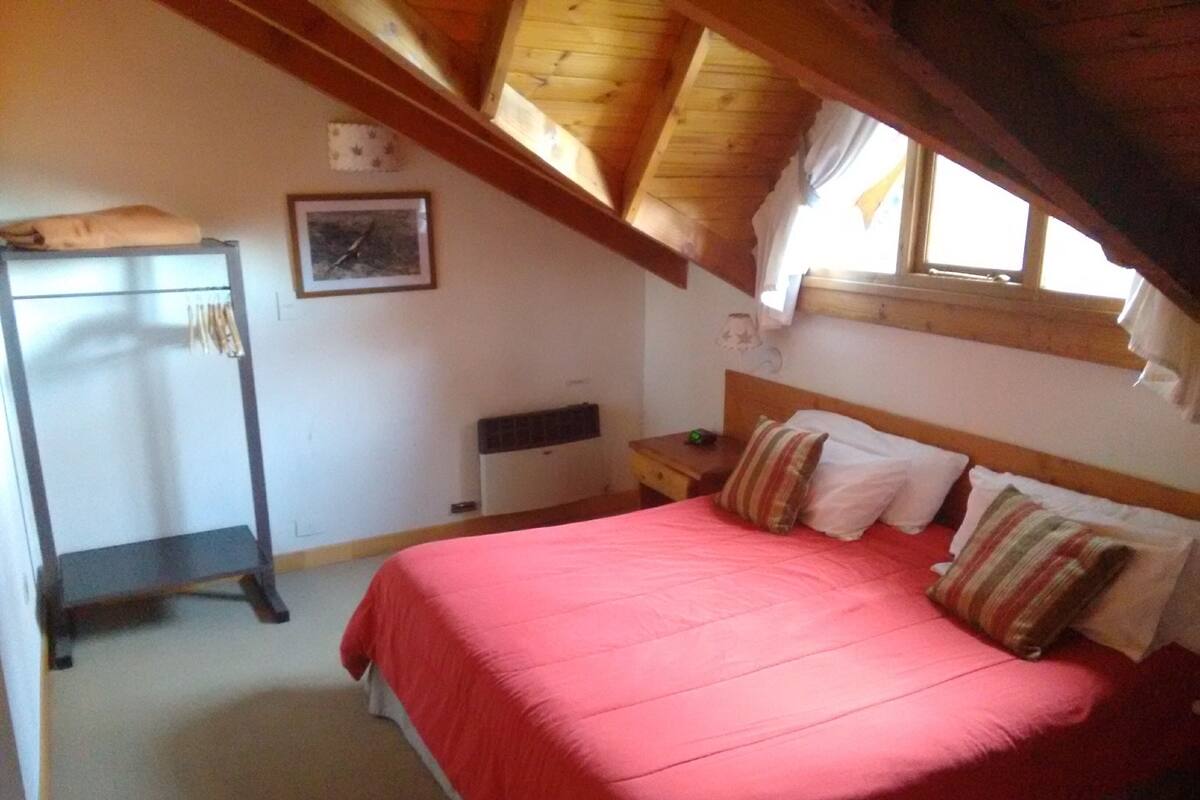 Ein Schlafzimmer mit Bett, Kissen, einem Nachttisch, einem Fenster und einer Holzbalkendecke.
