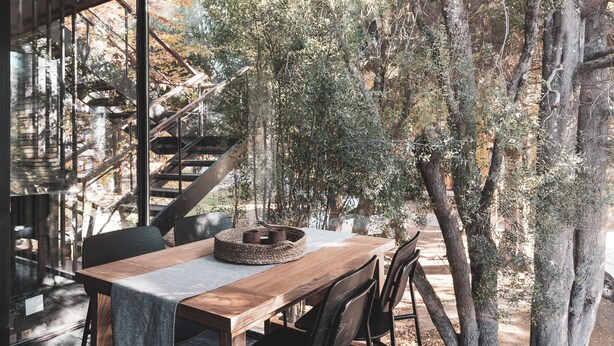 A wooden table with chairs, a woven basket, and a view of a forested area with a staircase.