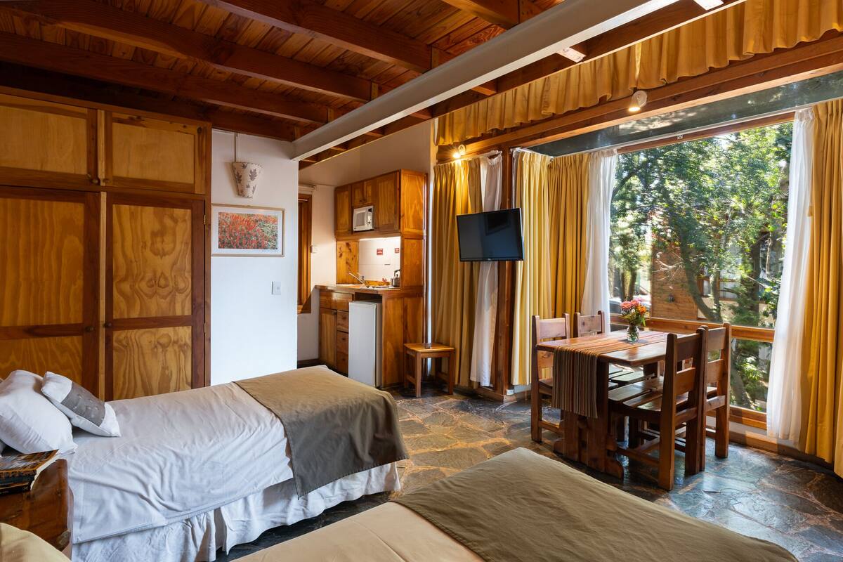 Ein gemütliches Zimmer mit einem Bett, einem kleinen Tisch mit Stühlen, einem Fernseher und einem Fenster mit Vorhängen.