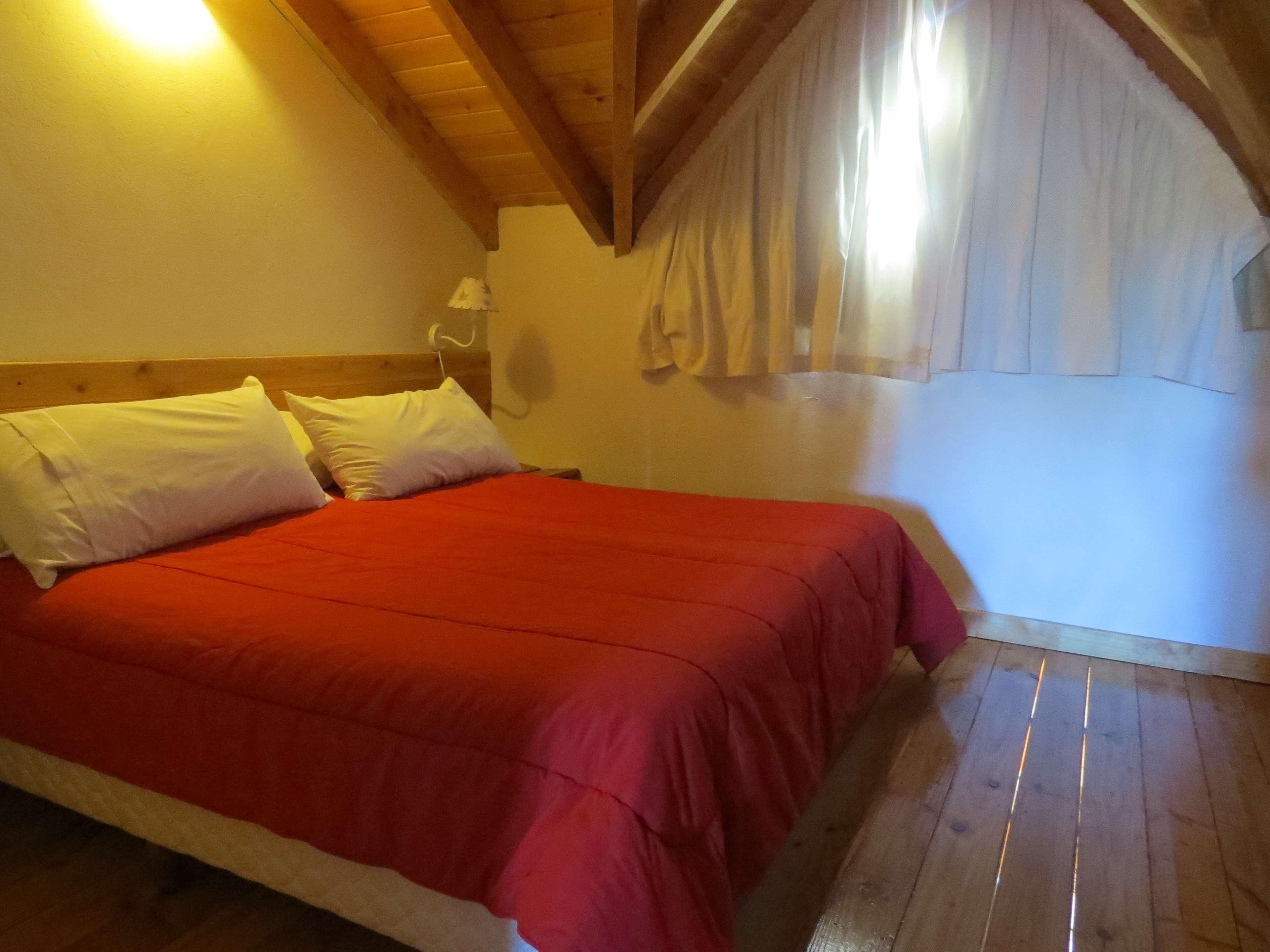 Een kamer met een bed, houten balken in het plafond, een ventilator aan de muur en een raam met gordijnen.