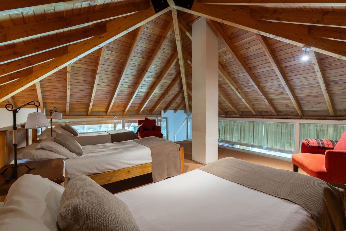 A high-ceilinged room with wooden beams, two beds, a red chair, and a window with curtains.