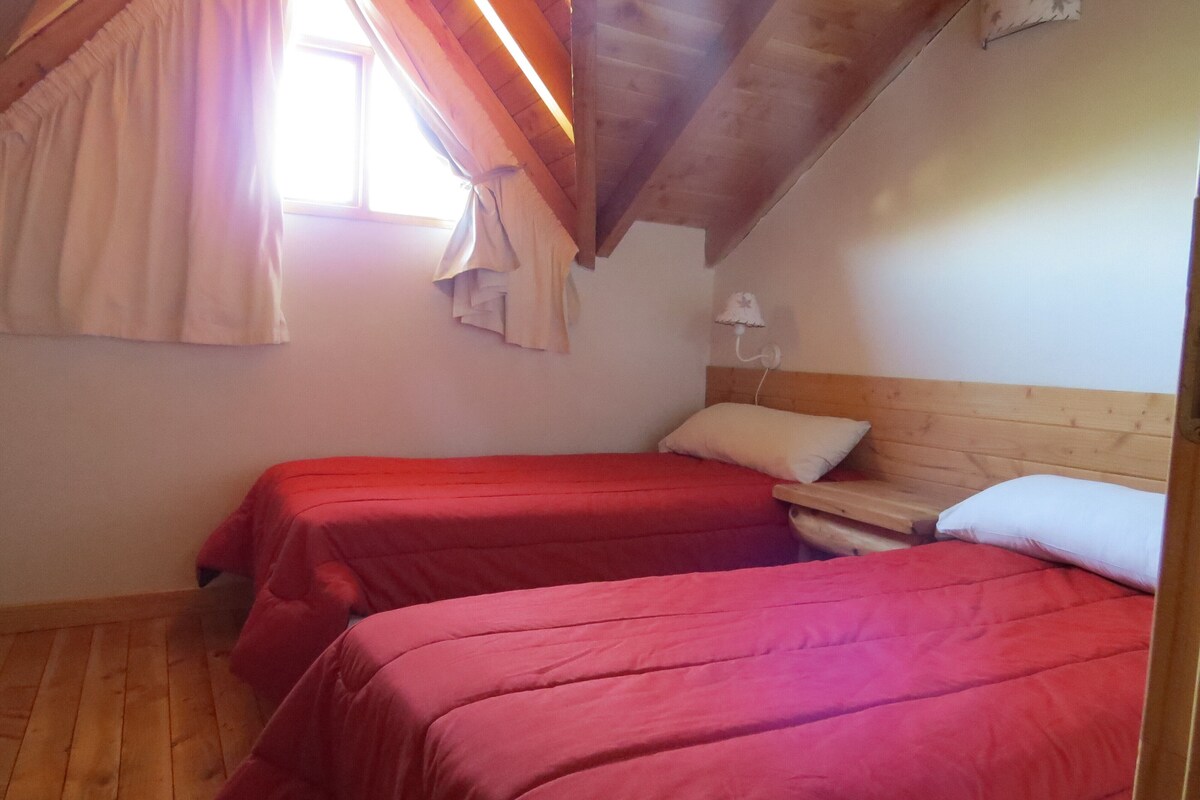 Une chambre avec deux lits, un plancher en bois, un plafond en pente et une fenêtre avec des rideaux.