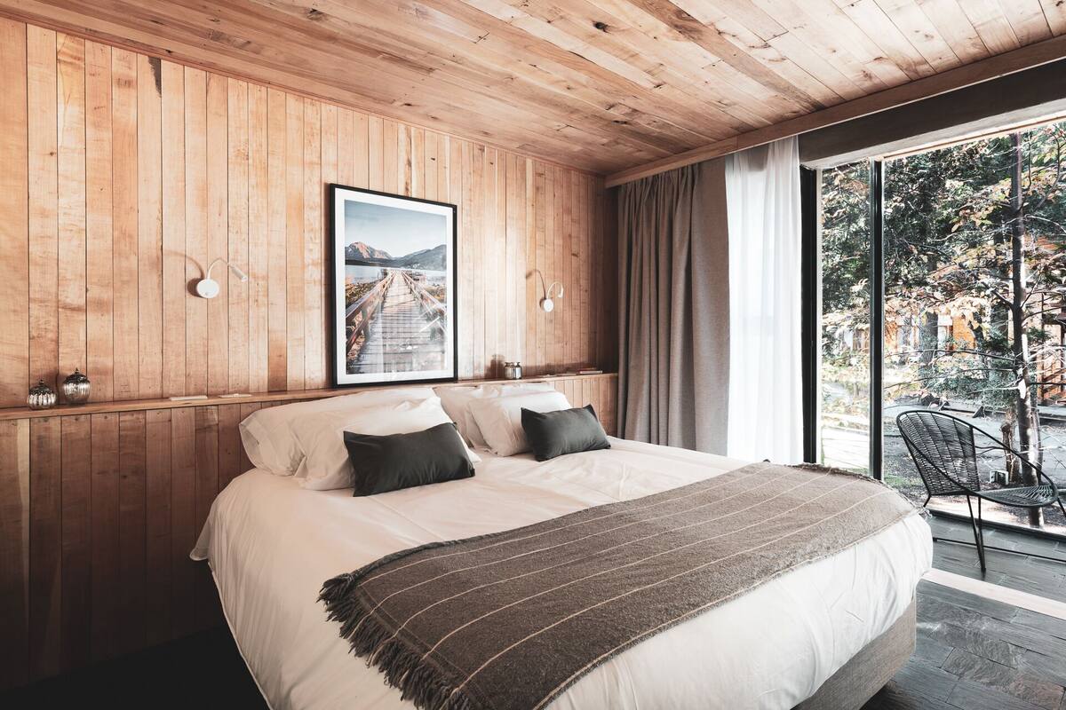Ein Schlafzimmer mit einem großen Bett, Holzwänden, einem gerahmten Bild und Blick auf einen Garten durch ein großes Fenster.