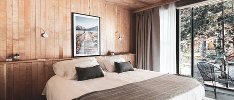 A bedroom with a large bed, wooden walls, a framed picture, and a view of a garden through a large window.
