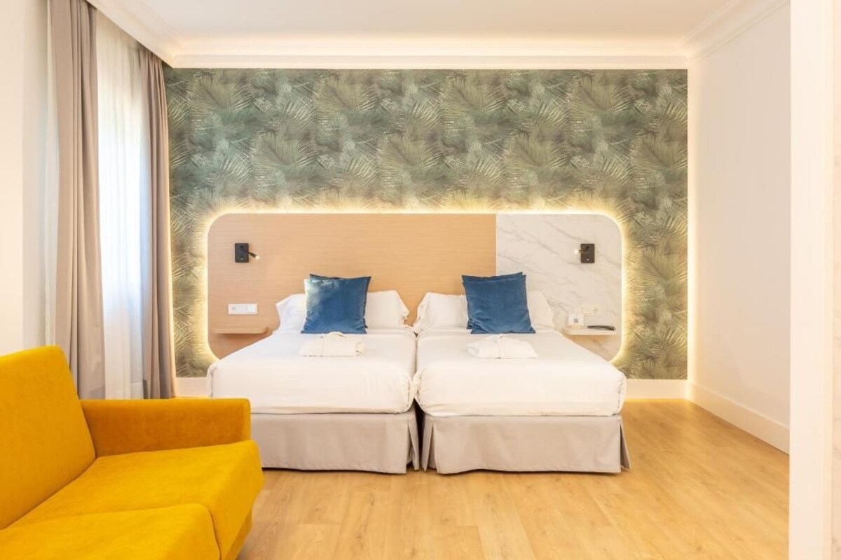 Habitación de hotel con cabecera dividida, dos camas con sábanas blancas y almohadas azules, un sillón amarillo y suelo de madera.