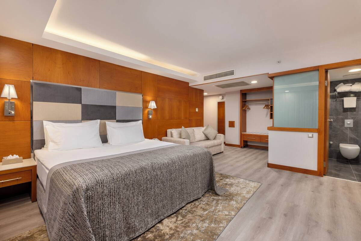A modern hotel room with a large bed, a sofa, a walk-in closet, and a bathroom with a toilet and sink.
