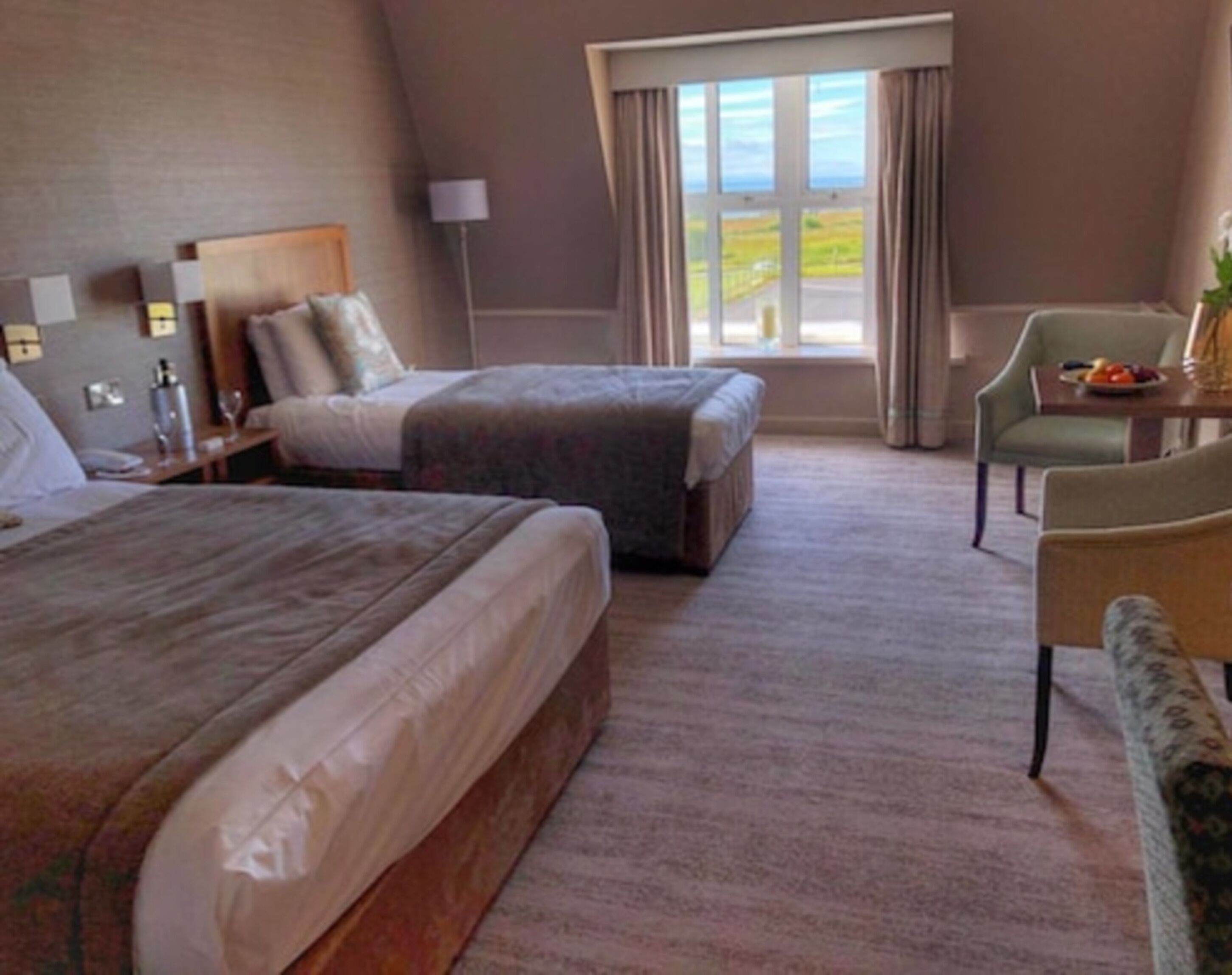 A hotel room with two beds, a sitting area with a chair and table, a lamp, and a window with a view.