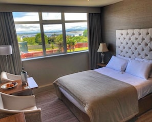 Une chambre d  ’hôtel avec un grand lit, un bureau avec une chaise, une lampe et une vue sur un paysage par la fenêtre.